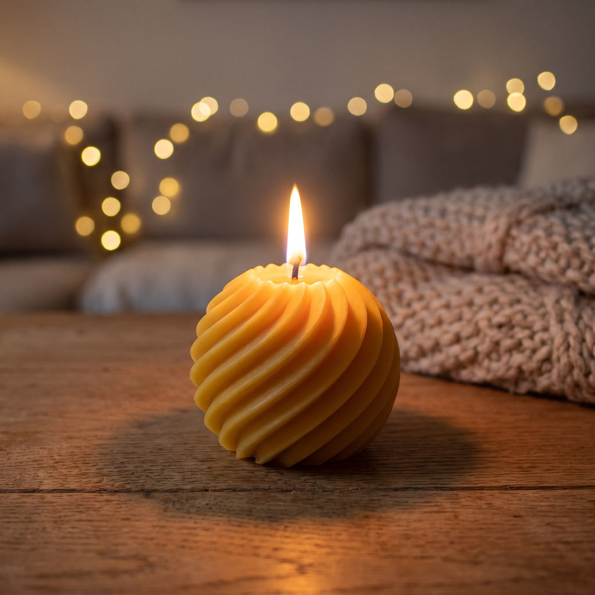 Spiralkugel aus Bienenwachs brennt abends auf einem Holztisch, im Hintergrund Lichterkette und Strickdecke