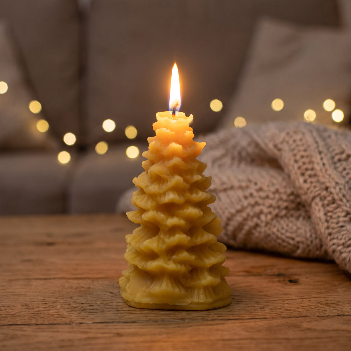 Kleine Tannenbaum-Kerze aus Bienenwachs brennt abends auf einem Holztisch, im Hintergrund Lichterkette und Strickdecke