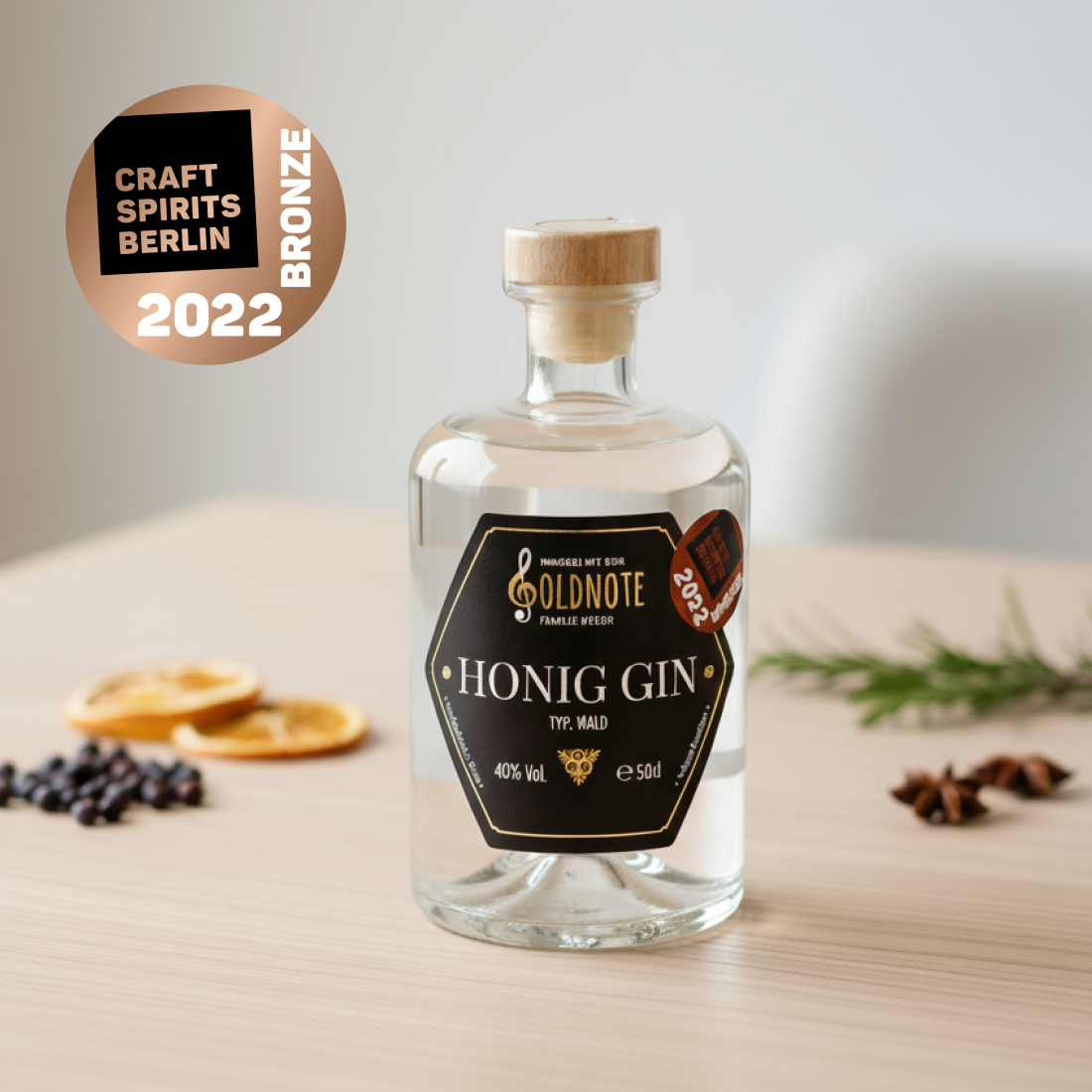 Flasche Goldnote Honig Gin mit Bronze-Auszeichnung der Craft Spirits Berlin 2022 auf Holztisch, dekoriert mit Wacholderbeeren und Orangenscheiben.